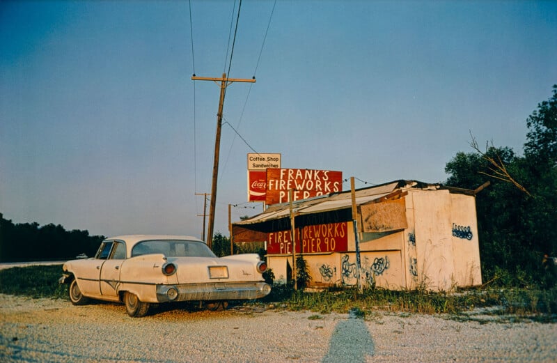 An old, dusty white car is parked in front of a small, rundown roadside stand with signs reading "Frank's Fireworks Pier 90." Power lines and trees line the background under a clear sky at sunset.