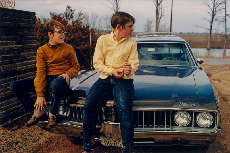 Two boys sit on the hood of a vintage blue Oldsmobile car parked outdoors. One boy wears glasses and a brown sweater; the other wears a yellow shirt. Bare trees and a road are visible in the background.