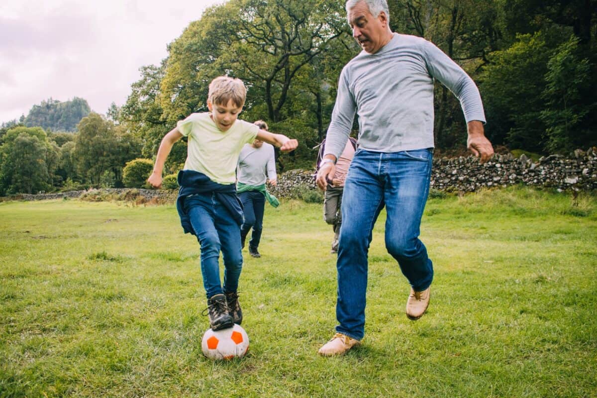 Three generation family are playing football together in a field. There are two boys, their father and their grandfather.