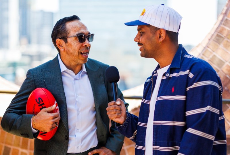 Two men engage in a conversation outdoors, one holding an Australian rules football. The man on the left wears sunglasses and a suit, while the man on the right, dressed casually in a striped jacket and cap, holds a microphone. The background features a blurred cityscape, suggesting a lively urban setting.