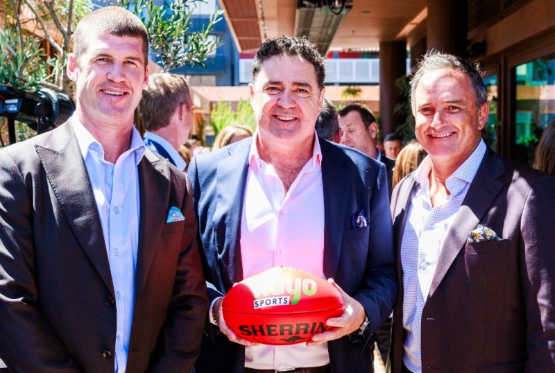 Three men pose together at a lively outdoor event, each dressed in smart casual attire. The central figure holds a Sherrin Australian Rules football, while the background features a crowd enjoying the sunny atmosphere. The image captures the essence of camaraderie and celebration within the Australian sporting community.