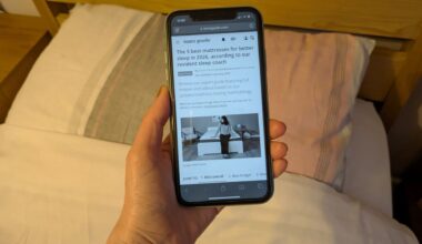 This image shows a hand holding an iPhone showing the Tom's Guide best mattress page in grayscale. In the background is a bed and pillows
