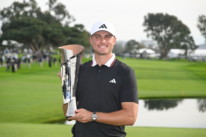 Golfer Ludvig Aberg, in a black Adidas polo and white hat, holds the trophy up and smiles on the course at the Genesis Invitational 2026.