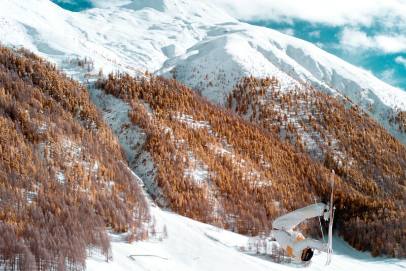 A snowboarder in white gear performs a mid-air flip against a backdrop of snow-covered mountains and golden-brown trees under a blue sky.