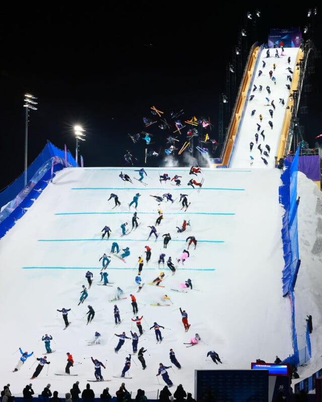 Skiers perform tricks and jumps as they descend a brightly lit snowy ramp at night, with multiple skiers in mid-air and others skiing down in parallel lanes, watched by spectators below.