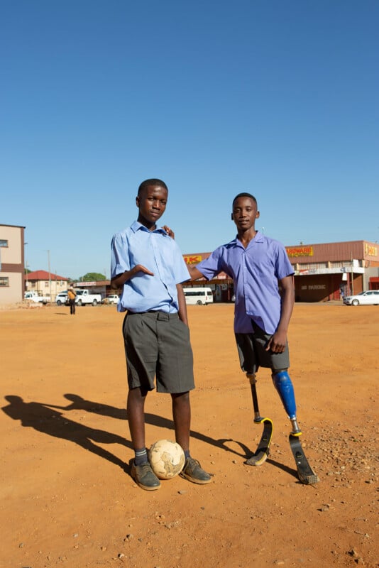 Two boys stand on a dusty field with a soccer ball. Both wear school uniforms; one has a prosthetic arm, and the other has two prosthetic legs. Buildings and cars are visible in the background under a clear blue sky.