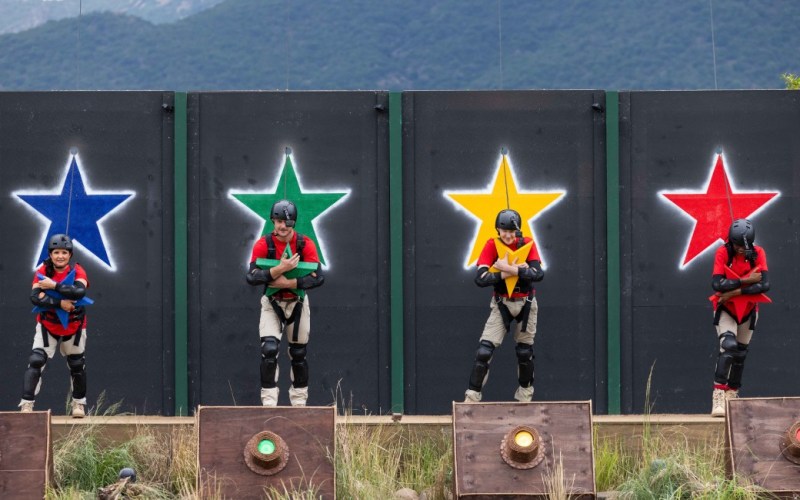 Four competitors stand in front of brightly coloured star-shaped targets on a challenge course, wearing protective gear and harnesses. Each participant is holding a differently coloured object, with vibrant backgrounds of blue, green, yellow, and red stars. The setting features a natural landscape, hinting at an outdoor competition or reality TV show.