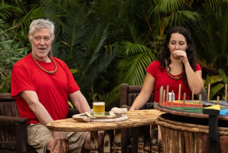 Two people seated at a wooden table in a lush outdoor setting, both wearing red shirts. The older man has grey hair and is looking ahead with a surprised expression, while the younger woman appears contemplative, covering her mouth. A glass of yellow drink is on the table, surrounded by natural greenery.