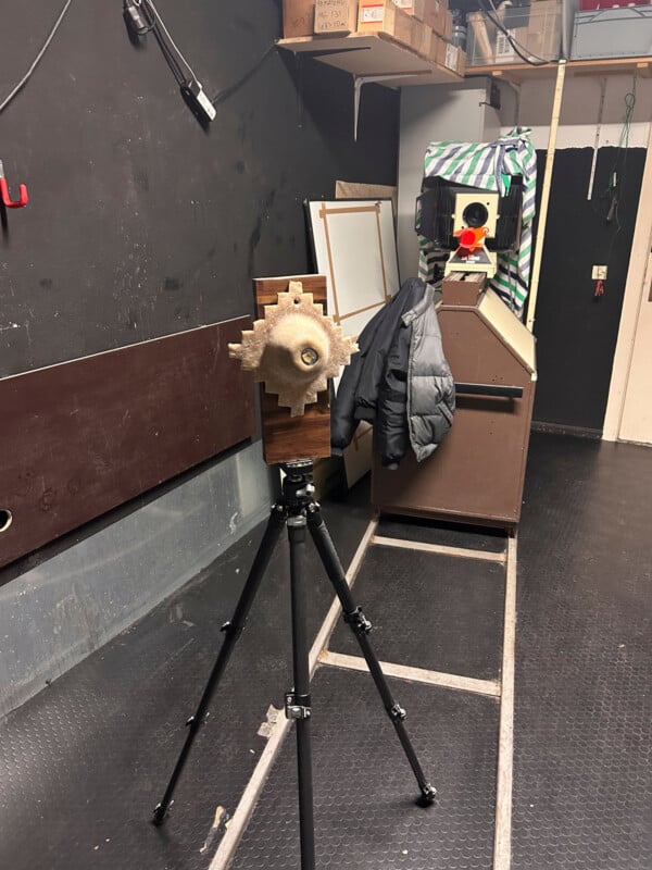 A wooden target with a large bullet hole mounted on a tripod stands in the foreground of an indoor shooting range. In the background, there’s a padded enclosure, a black jacket, and various storage items.