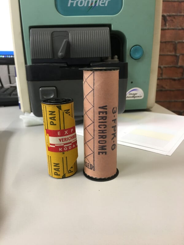Two vintage film rolls, one yellow labeled "PAN" and the other larger and tan labeled "VERICHROME," stand upright on a desk in front of an office machine with a brick wall in the background.