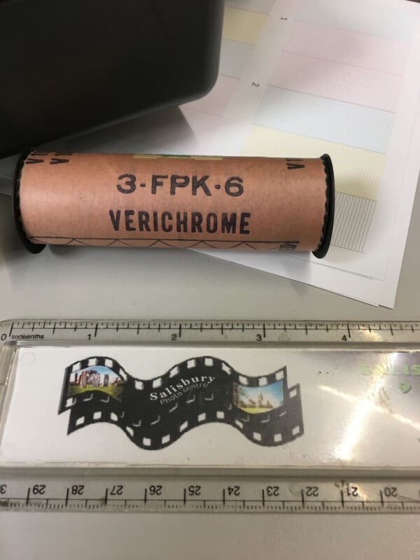 A vintage Verichrome film roll is on a desk near a clear ruler with a filmstrip design labeled "Salisbury." There are also some color test printouts and part of a black plastic object visible in the background.