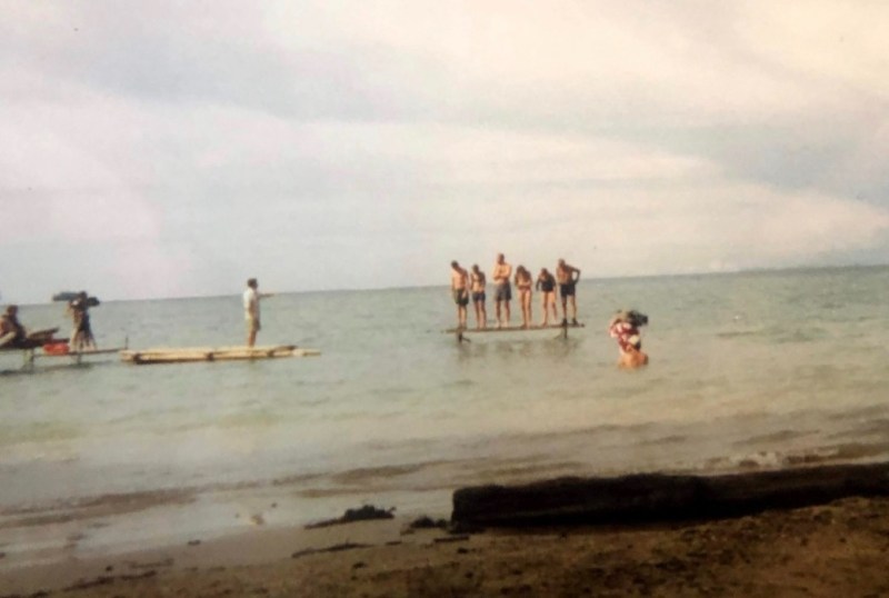 Group of people enjoying a beach day in Australia, with some standing on a wooden platform in the water while others swim nearby. Cloudy sky and calm sea create a relaxed atmosphere.