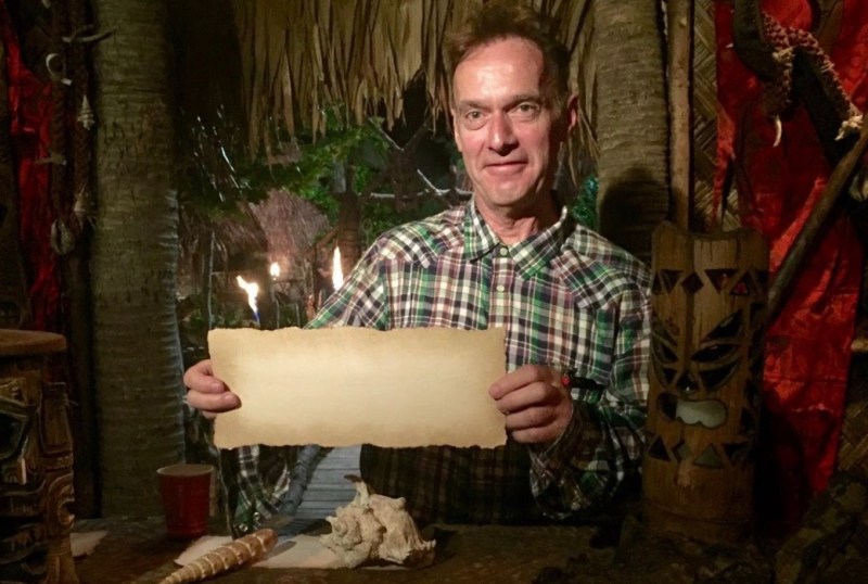 Man holding an old parchment in a tropical-themed setting, surrounded by tiki decor and palm trees, with torches in the background.