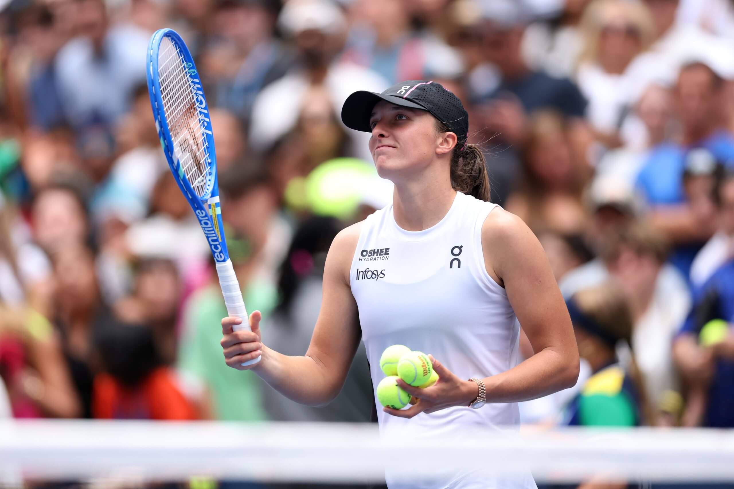 Iga Świątek raises her racket in celebration holding three tennis balls in her left hand, with a crowd blurred behind her in the background.