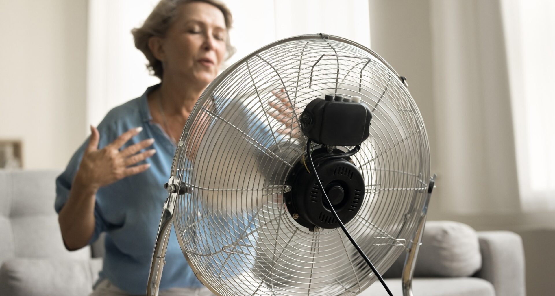 Close up electric ventilator with propeller cooling air blowing to senior older lady suffering from overheating, hypoxia, sitting on sofa with closed eyes, flying hair in blurred background