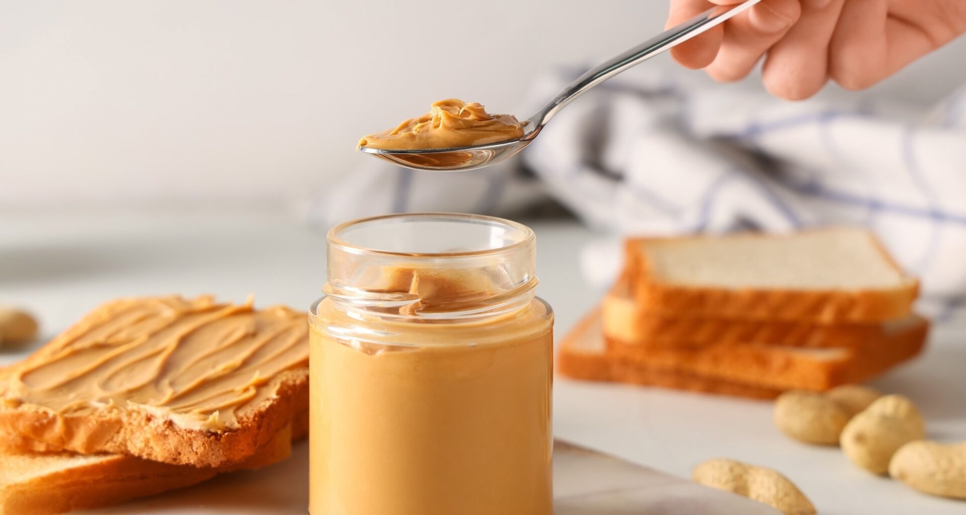 Female hand with jar of tasty peanut butter, spoon and toasts on white background