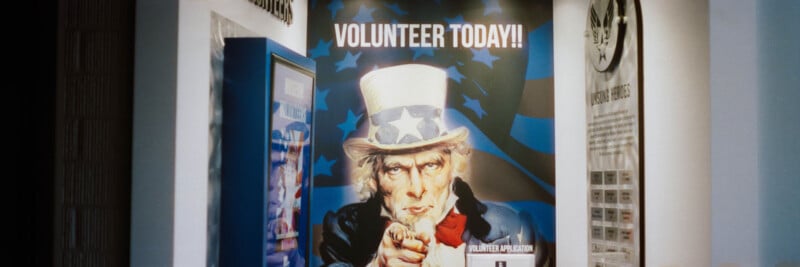 A poster featuring Uncle Sam pointing outward with the text "VOLUNTEER TODAY!!" in bold letters above, displayed on a wall in a hallway with other informational displays nearby.