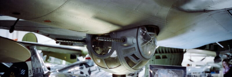 Close-up view of a World War II-era bomber’s ball turret with twin machine guns, mounted under the aircraft. Vintage planes and museum displays are visible in the background.