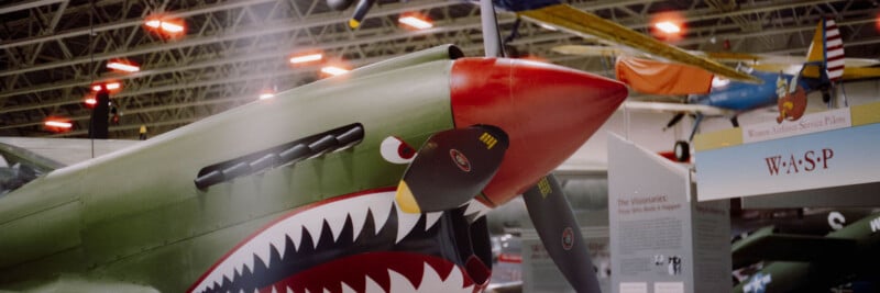 A close-up of a vintage military aircraft with a shark mouth painted on its nose, displayed in an aviation museum with other planes and a WASP sign visible in the background.