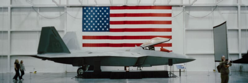 A gray fighter jet displayed indoors in front of a large American flag, with a few people walking and standing nearby in a spacious, brightly lit hangar.