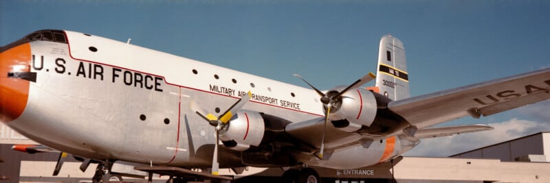 A large silver U.S. Air Force Military Air Transport Service cargo plane with four propellers is parked outside a building under a blue sky.