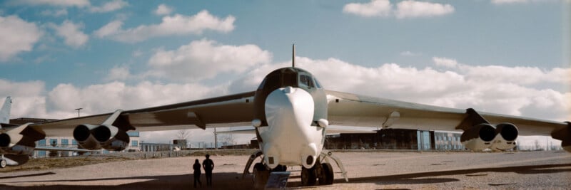 A large military aircraft, likely a B-52 bomber, is displayed outdoors on a gravel area under a partly cloudy sky. Two people stand near the nose of the plane, highlighting its size.