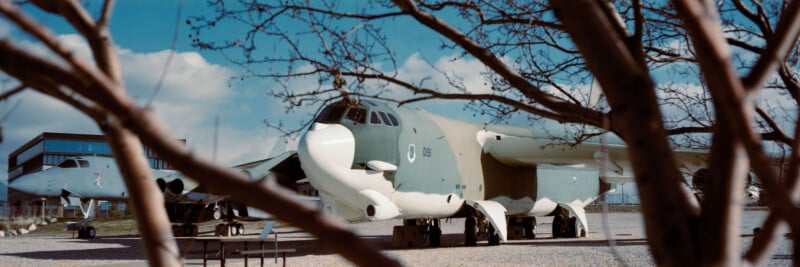 A large military aircraft with camouflage paint is parked on gravel outdoors, partially obscured by leafless tree branches. Another similar plane and a building are visible in the background under a blue sky with clouds.
