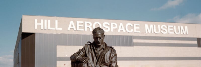 A bronze statue of a seated figure is in front of a large building with "HILL AEROSPACE MUSEUM" written on its facade under a blue sky.