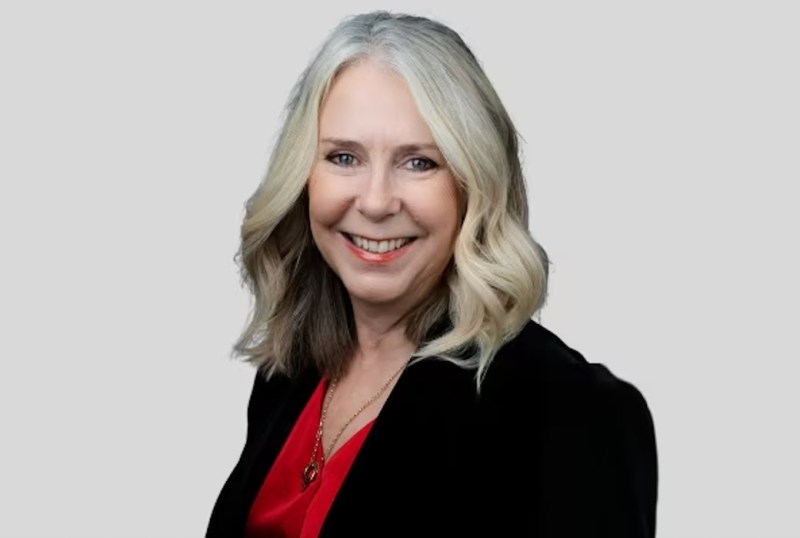 Professional headshot of a woman with shoulder-length blonde hair, wearing a black blazer and a red blouse, smiling against a neutral grey background.