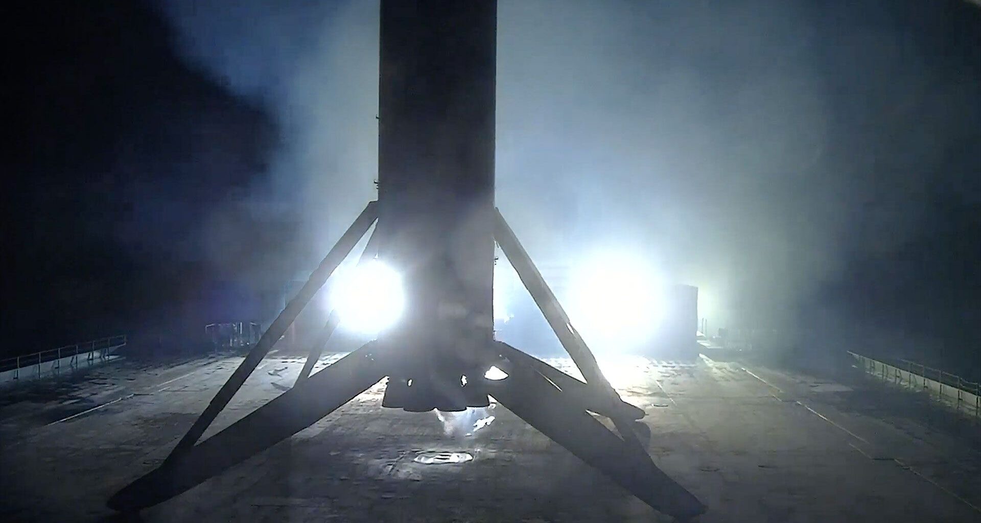 the first stage of a rocket is seen lit at night in silhouette standing on its four landing legs atop a sea-based droneship