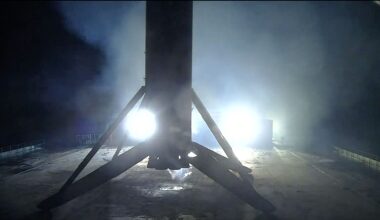 the first stage of a rocket is seen lit at night in silhouette standing on its four landing legs atop a sea-based droneship