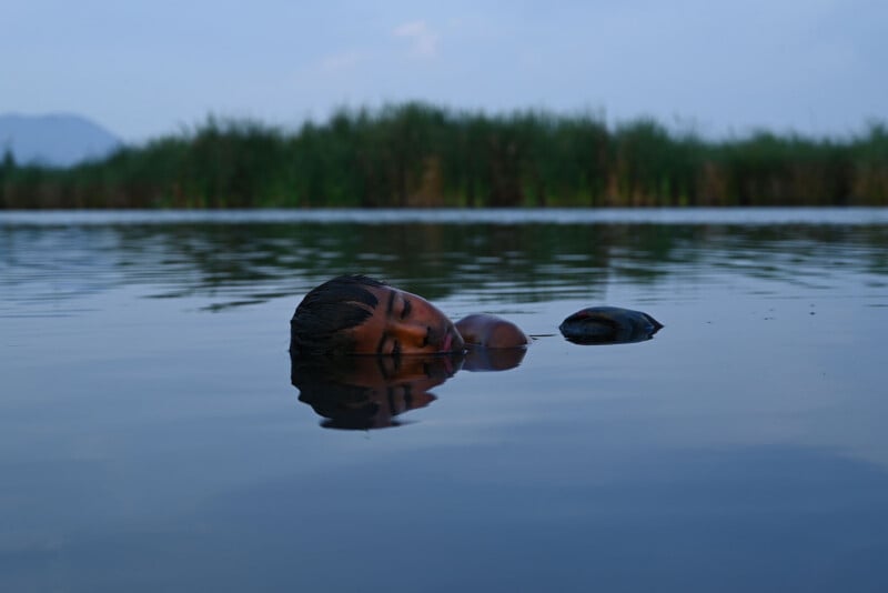 A child floats peacefully on their back in calm water, with eyes closed and face partially submerged, surrounded by greenery and distant mountains under a blue sky.