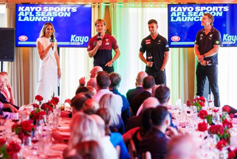 Kayo Sports AFL season launch event featuring a presenter and three players speaking to an audience, with a backdrop displaying the Kayo Sports logo. The venue is decorated with red roses, and attendees are seated at tables enjoying the event.