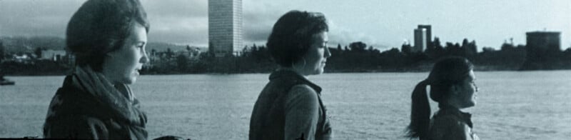 Three women in profile stand near a body of water, facing right. City buildings and trees are visible in the distant background under a cloudy sky. The image has a vintage, black-and-white appearance.