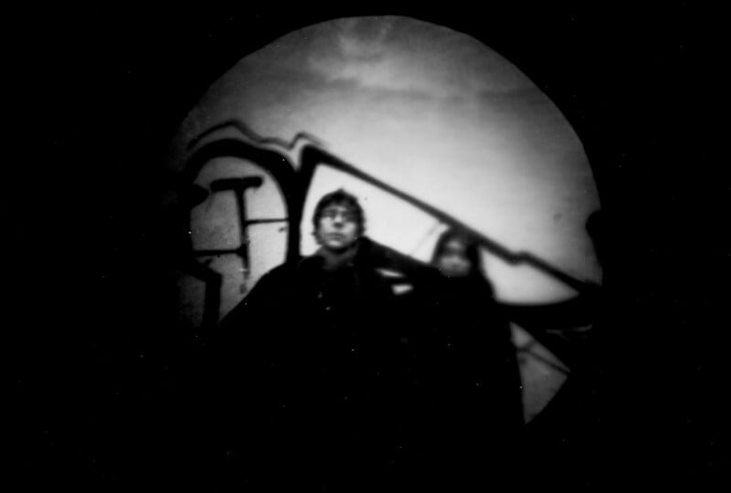 A blurry, black-and-white photo shows two people standing in front of a wall with graffiti. The image is vignetted, with the figures and background partially obscured by shadows and darkness.