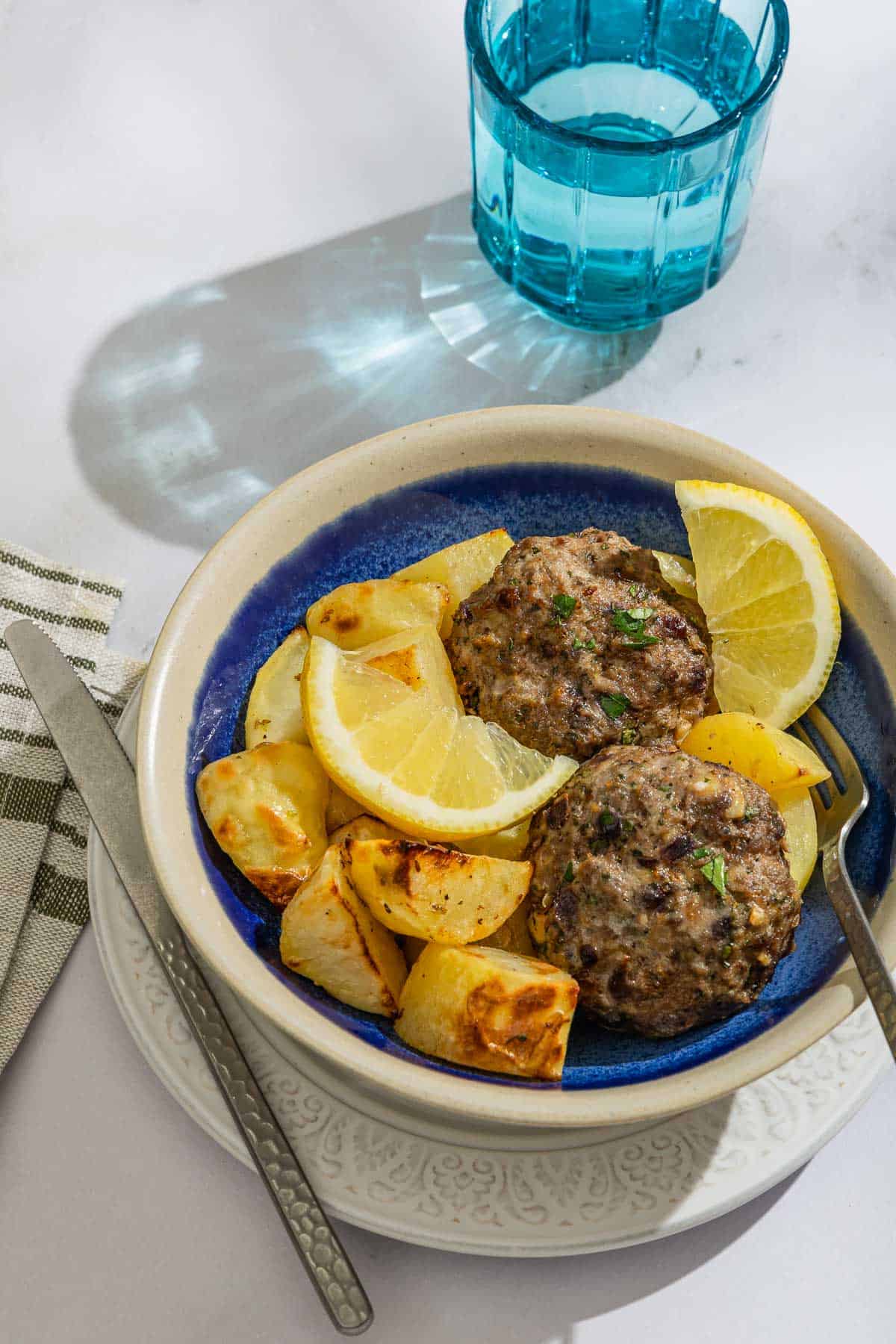 Bifteki with lemon potatoes and lemon wedges in a bowl with a fork. The bowl is on a plate with a knife and surrounded by a cloth napkin and a glass of water.