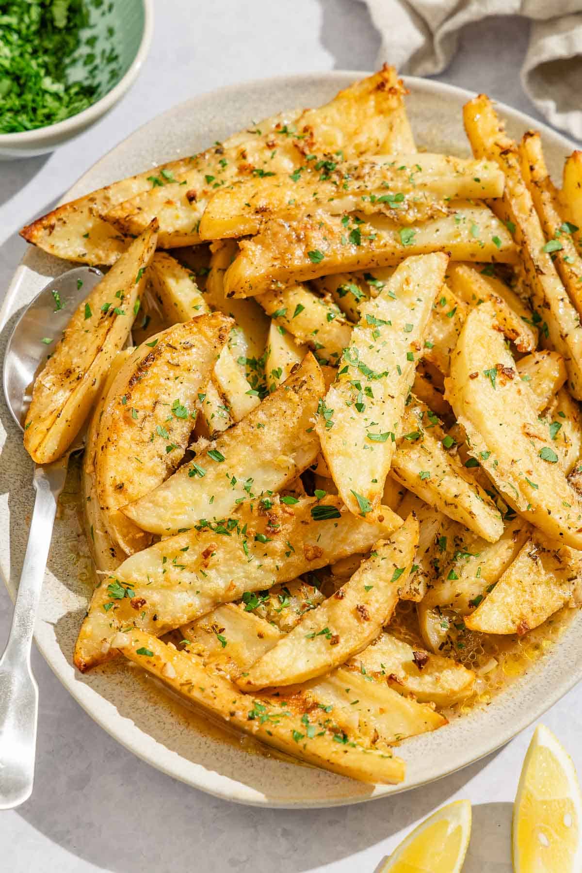 Greek lemon potatoes on a platter with a serving spoon. Next to this is a bowl of chopped parsley and 2 lemon wedges.