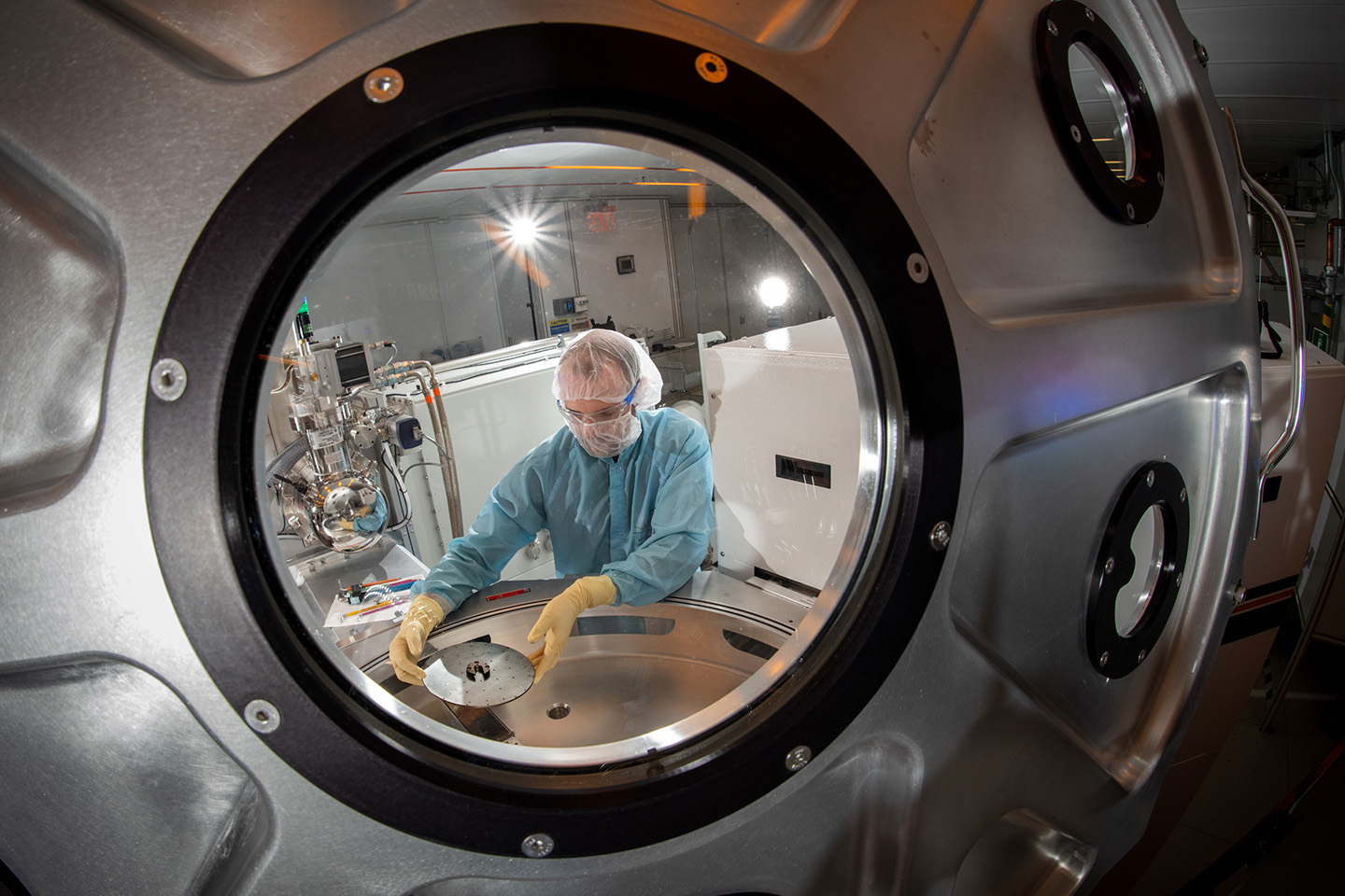 A researcher in light blue personal protective equipment adjusts a small metal disk within the cluster tool. The researcher is framed through a circular glass window on the experimental setup.