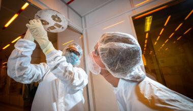 One researcher holds up a disk while another researcher peers at the underside. Both researchers are wearing white personal protective equipment and stand in front of orange reflective lab windows.