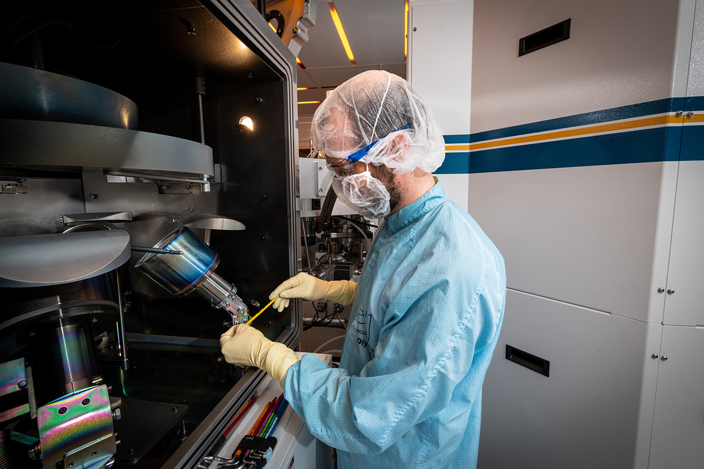 A researcher in light blue personal protective equipment stands to the right of a scientific setup and prepares a sample.