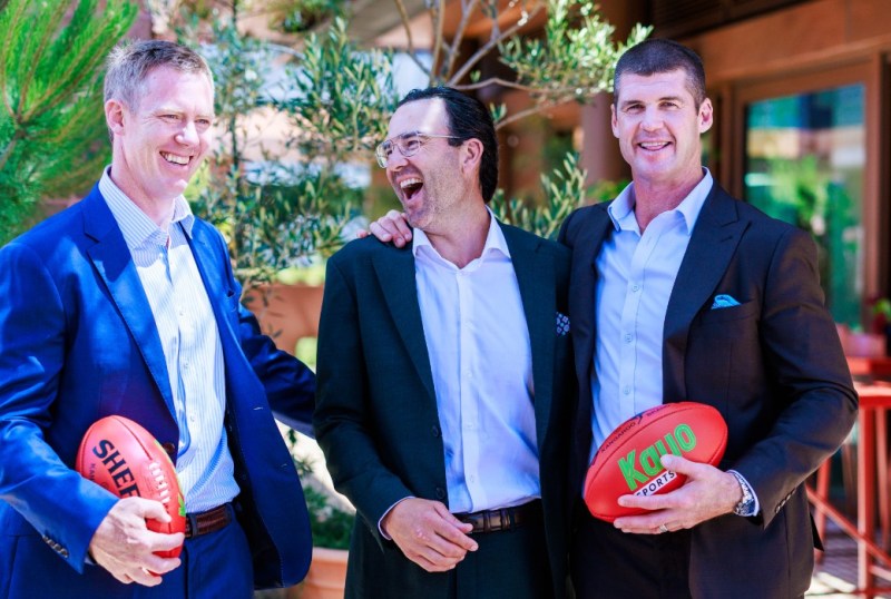 Three professional men laughing and holding Australian rules footballs outdoors, dressed in smart casual attire against a backdrop of greenery, showcasing a relaxed and engaging atmosphere.