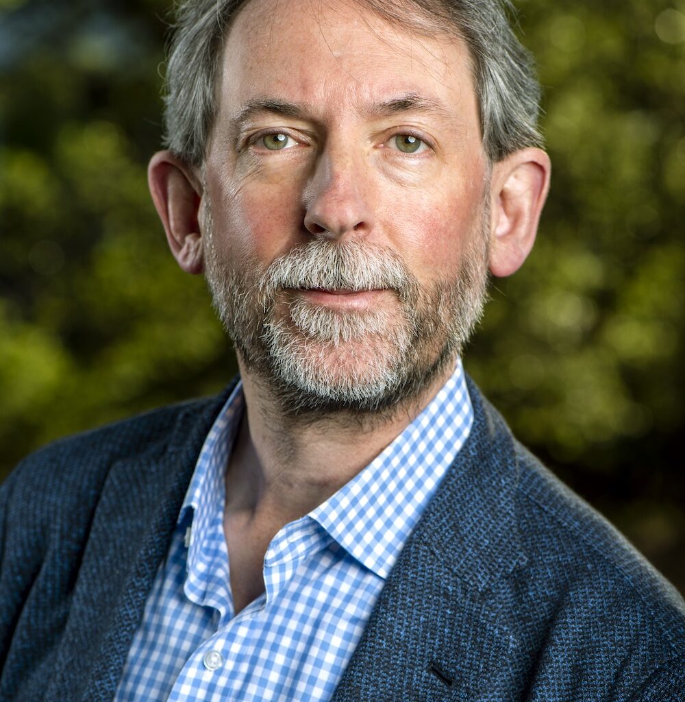 A man in a dark blue suit poses for a profile photo against a blurred green background