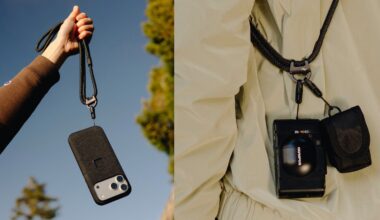 A hand holds a smartphone on a black lanyard against the sky, while another lanyard secures a camera and small pouch to a person in a light-colored jacket.