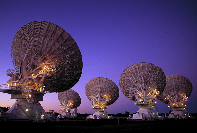 Five large white dish-shaped radio telescopes face away from the viewer, looking out to a purple dusk sky. The staircases leading up to the back of the telescopes are lit.