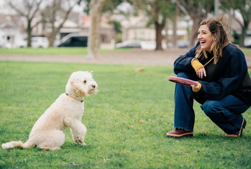 Woman playing with a fluffy white dog in a park, holding a frisbee and laughing while crouching on the grass.