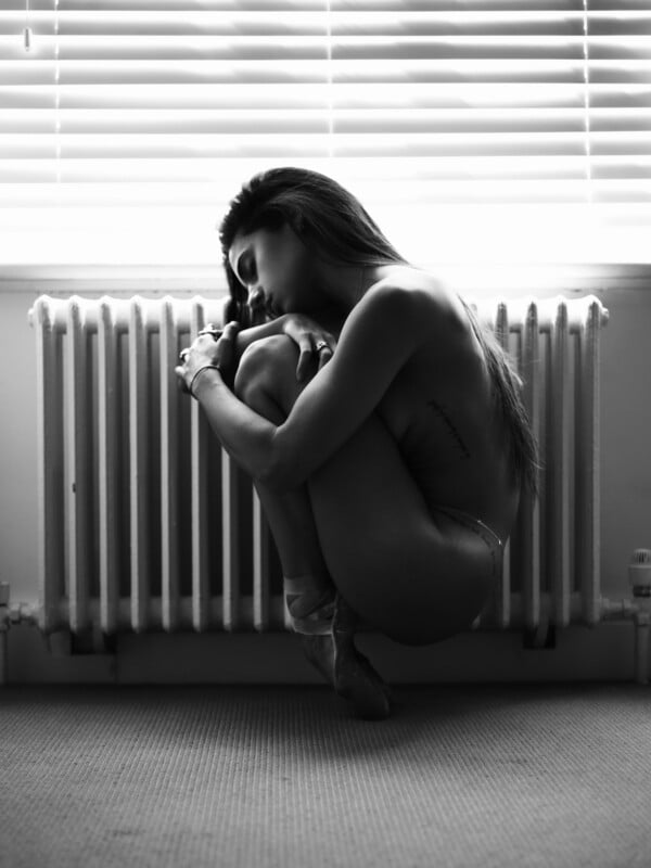 A woman sits nude in a fetal position on the floor in front of a radiator, lit by window blinds, in a black and white photograph.