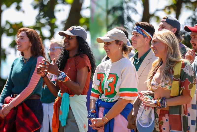 A diverse group of young adults cheer enthusiastically outdoors, dressed in colourful casual attire and accessories, showcasing a vibrant atmosphere during a lively event. The background features greenery, adding to the festive setting.