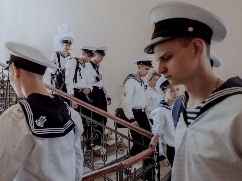 A group of young naval cadets in white uniforms and hats walk up and down a staircase inside a building with beige walls and iron railings. Some cadets are looking ahead while others look down or to the side.
