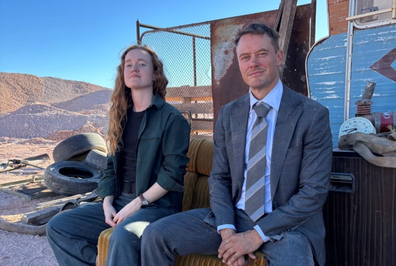 Two individuals seated on a vintage couch in a rugged outdoor setting, with a backdrop of rocky terrain and discarded tyres. The woman, dressed in a dark green outfit, has long wavy hair and appears contemplative. The man, in a formal grey suit and tie, sits confidently beside her, both facing the camera. The scene conveys a contrast between casual and formal attire amidst a desolate landscape.