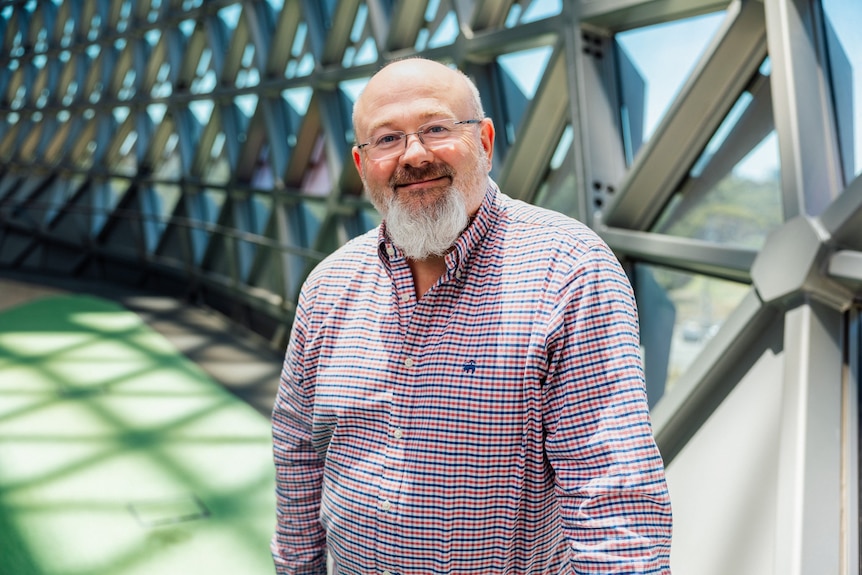 A man with a grey beard wearing a red and blue checked business shirt smiles.